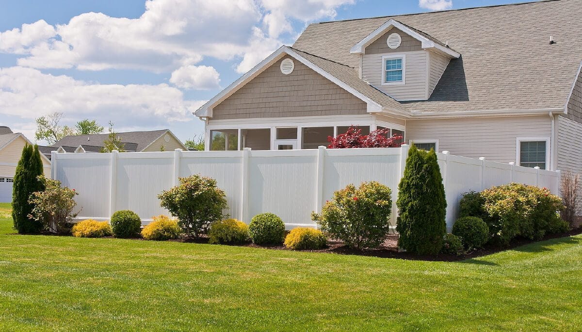 privacy fence cost A house with a neatly trimmed lawn, a white fence, and bushes and trees in the yard under a blue sky with white clouds.