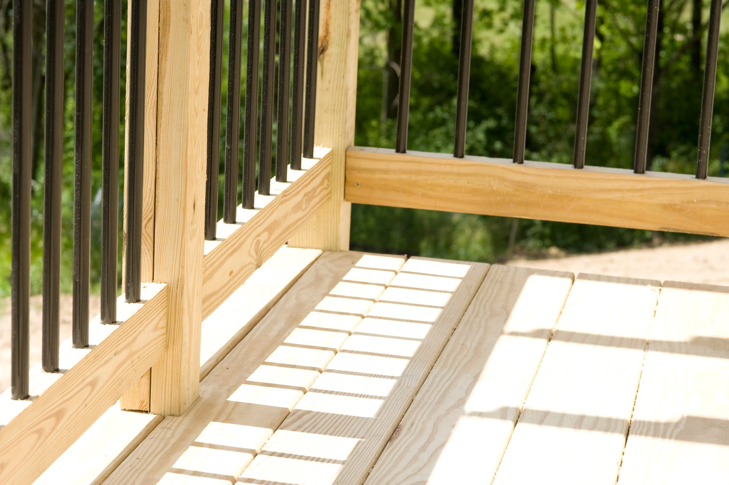 Sunlit wooden deck with black vertical railings casting shadows on the floor, surrounded by greenery.