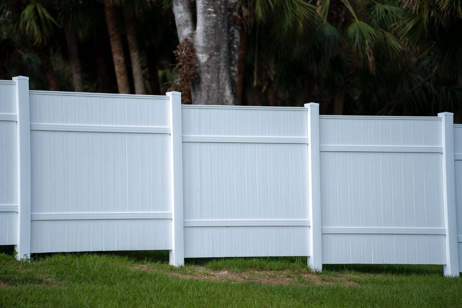White vinyl fence panels standing on green grass with trees and foliage in the background.