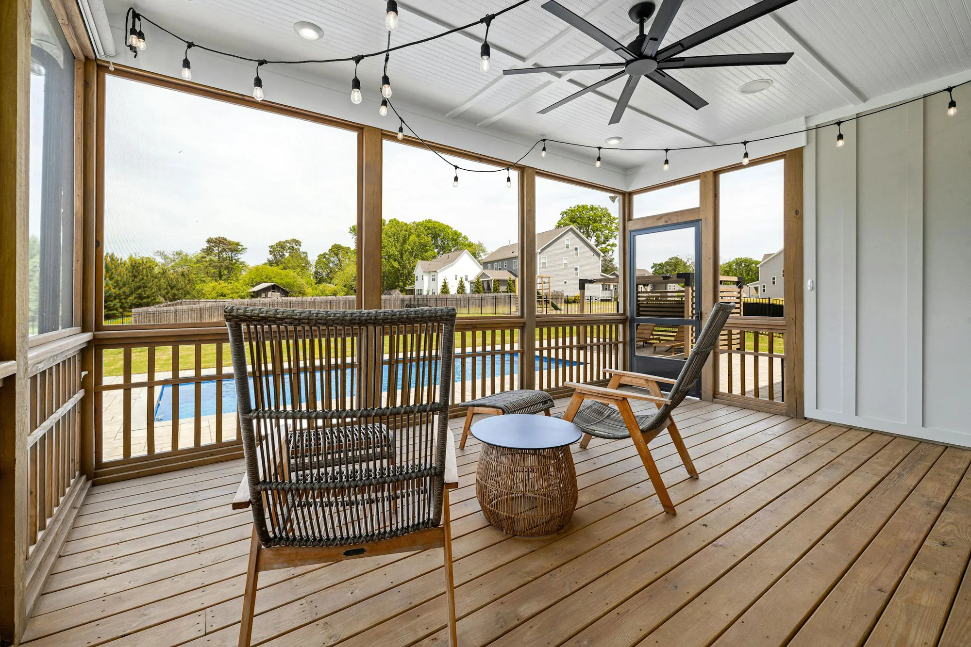 swimming-pool-behind-in-house Screened porch with wooden floor, chairs, string lights, and pool view in a suburban backyard.