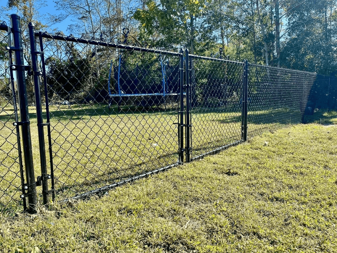 Black chain-link fence with a gate, bordering a grassy yard with trees in the background.