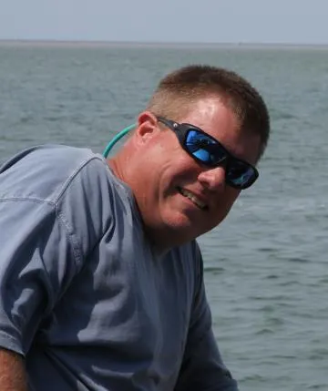 Man wearing sunglasses and a gray shirt smiles while sitting by the water on a sunny day.